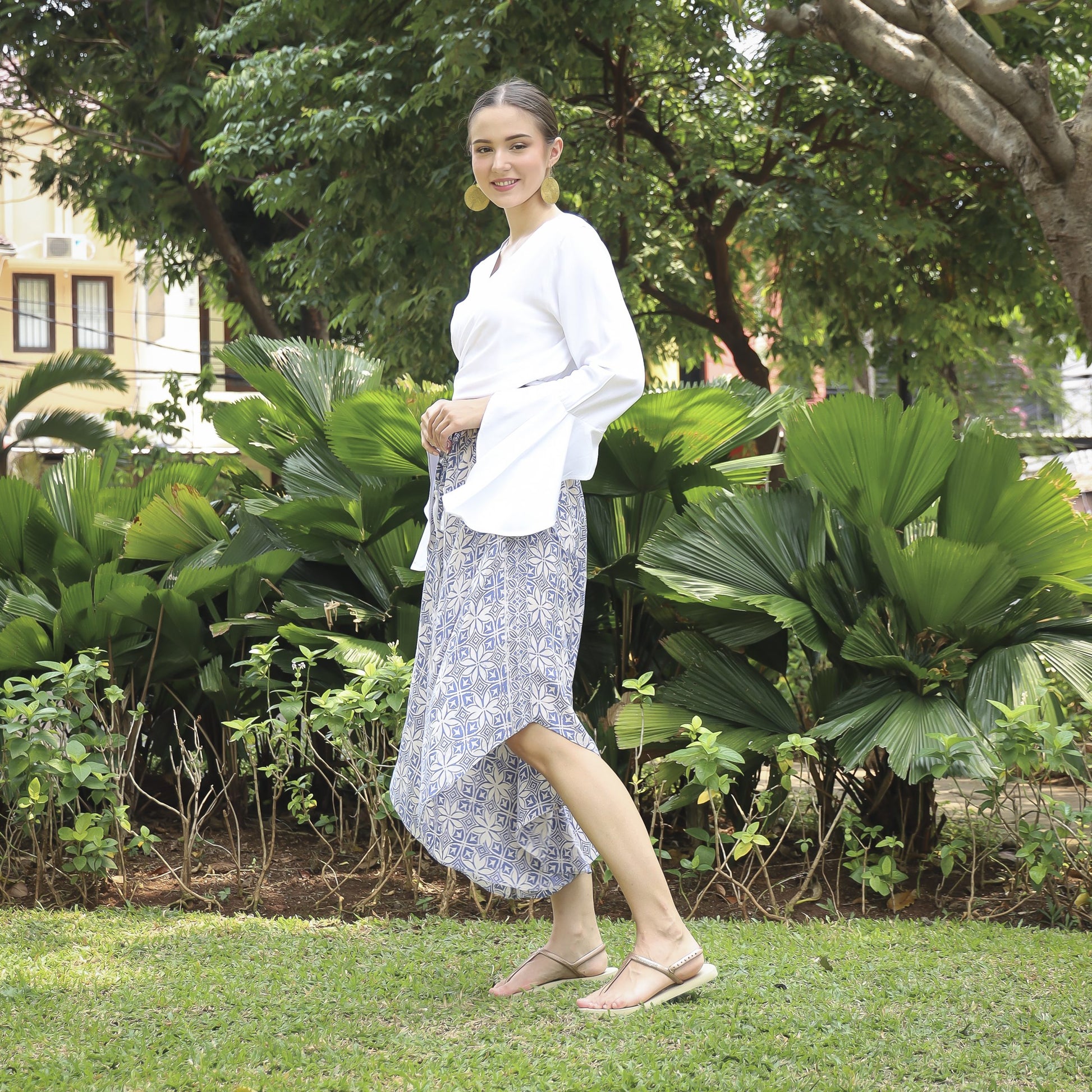 A woman in a white blouse and hand stamped batik pants smiles while standing on grass in a garden. Lush green plants and trees surround her, with colorful buildings in the background. She is wearing sandals and has her hair tied back.