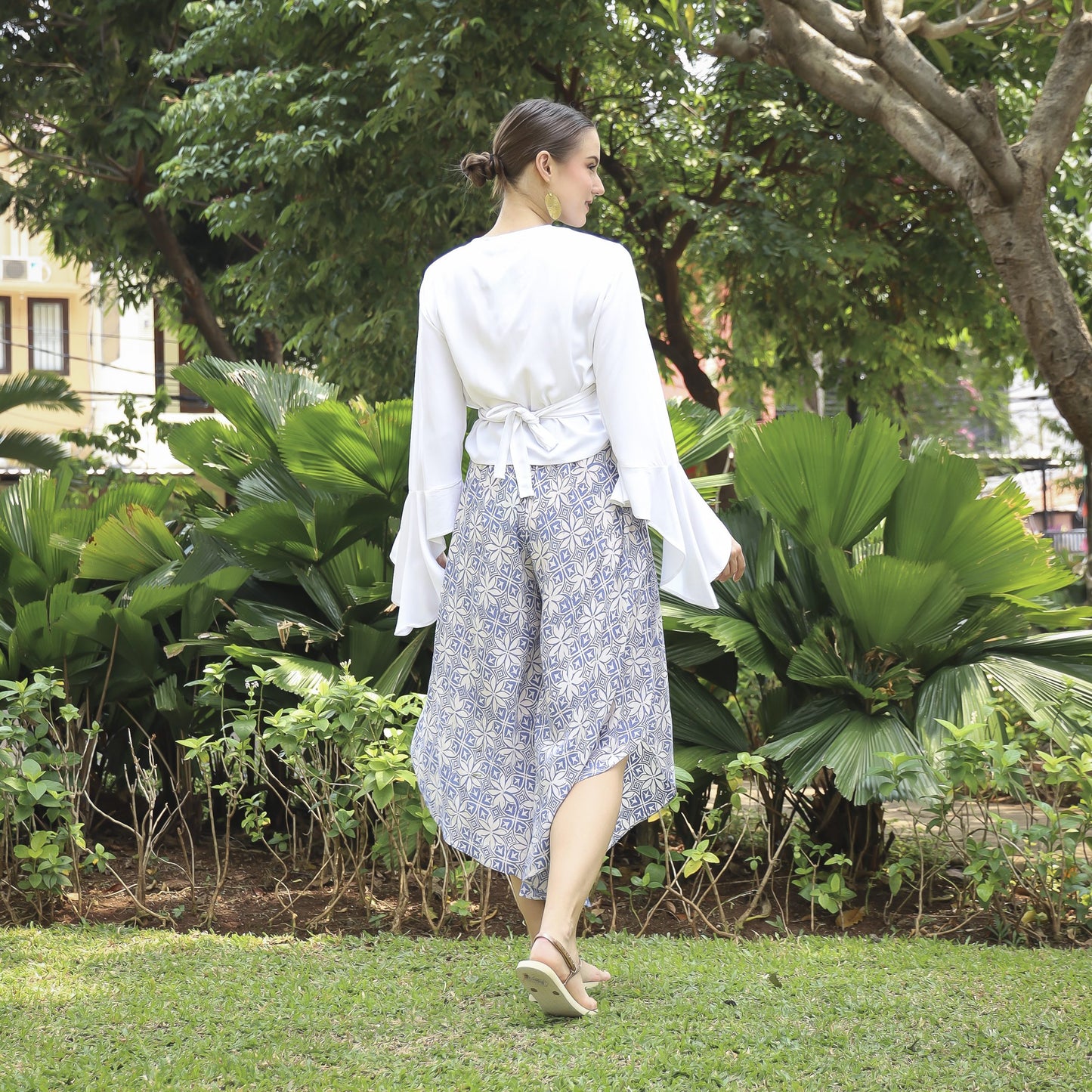 A woman wearing a white top and patterned skirt stands in a garden with lush greenery. She is facing away, with her hair in a bun, and wears sandals. The atmosphere is serene and natural.