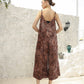 A woman stands outdoors on a light-colored patio against a textured white wall, wearing a patterned brown and black sleeveless dress. Her hair is styled in a bun. Nearby are a lantern and decorative jars. She faces away from the camera.
