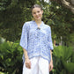 A woman stands outdoors in front of lush green foliage, wearing a comfortable batik top for women with blue and white patterns paired with white pants. She smiles softly, her hands relaxed at her sides.