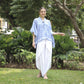 A person in a blue and white patterned shirt and long white skirt stands on grass in a lush garden, surrounded by trees and bushes. They are smiling and barefoot.
