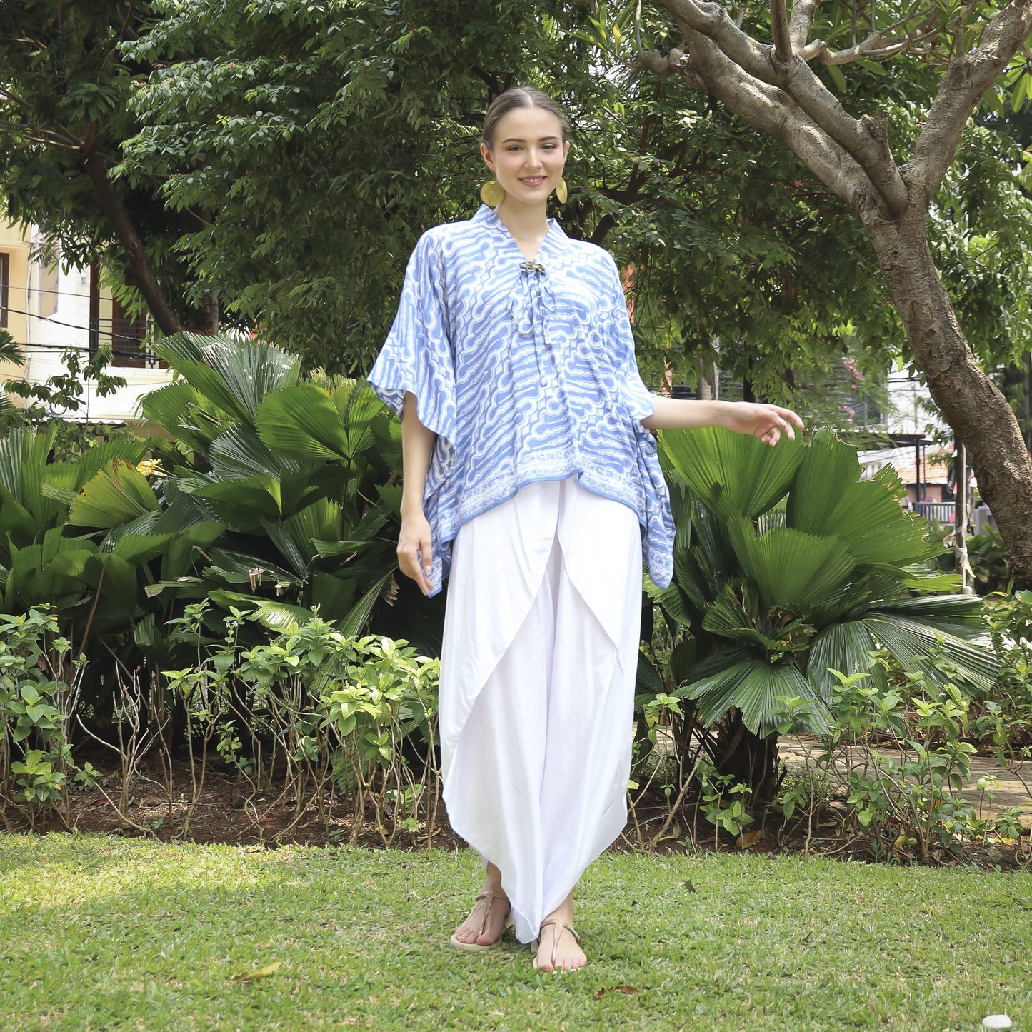 A person in a blue and white patterned shirt and long white skirt stands on grass in a lush garden, surrounded by trees and bushes. They are smiling and barefoot.
