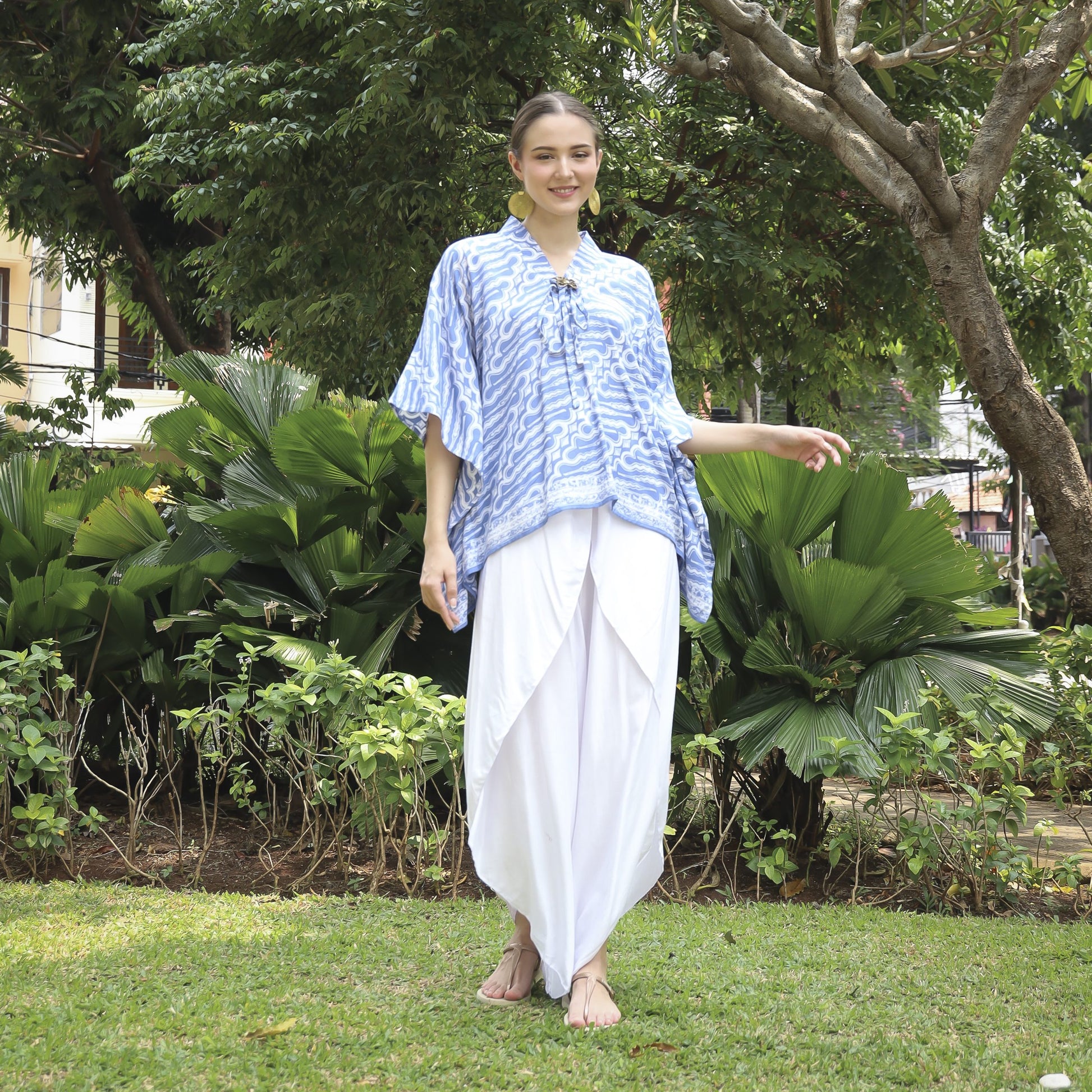 A person in a blue and white patterned shirt and long white skirt stands on grass in a lush garden, surrounded by trees and bushes. They are smiling and barefoot.