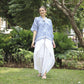 A person stands on grass in a park, adorned in a comfortable blue and white batik top for women and white flowing pants. They are barefoot and smiling, surrounded by the lush greenery of trees and bushes.