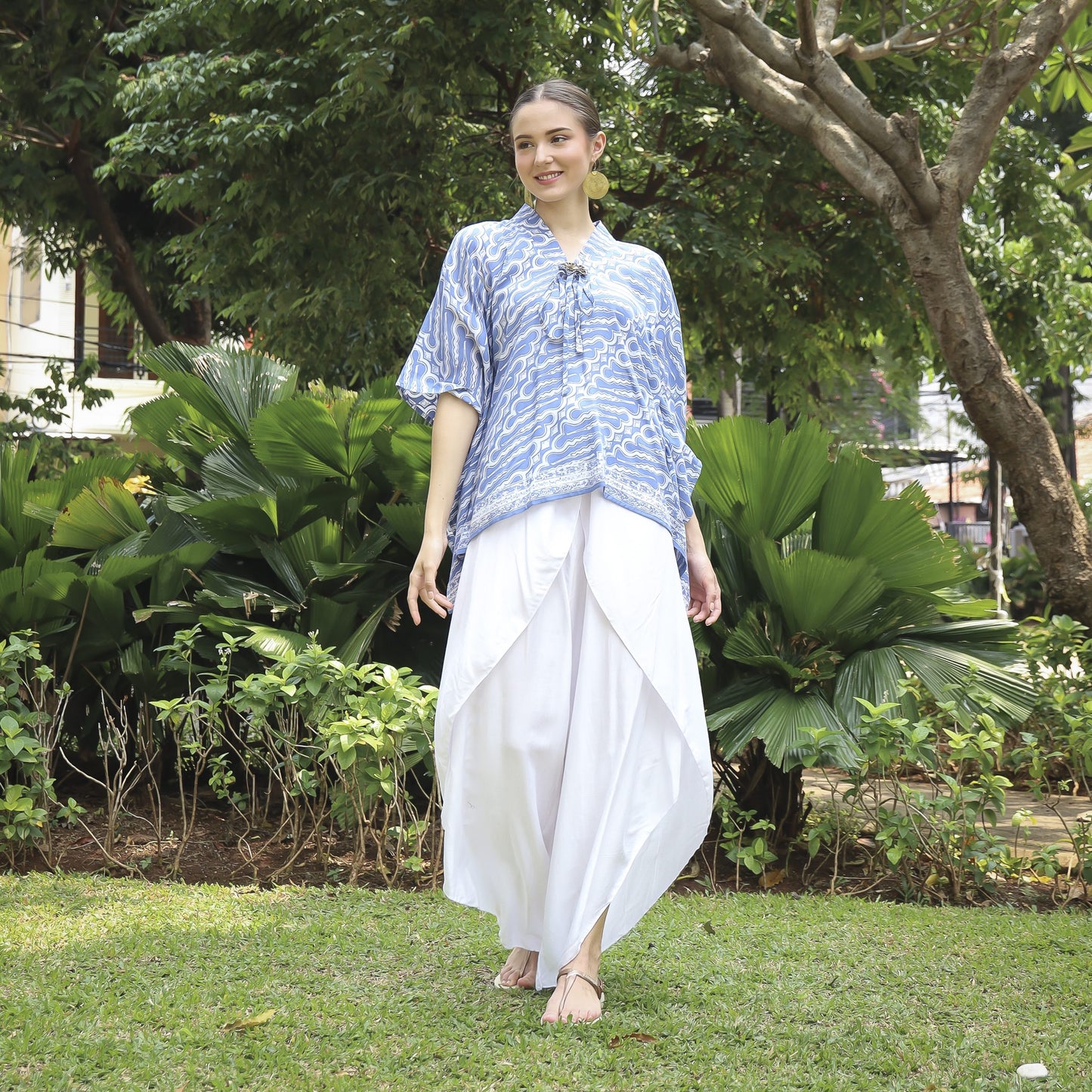A person stands on grass in a park, adorned in a comfortable blue and white batik top for women and white flowing pants. They are barefoot and smiling, surrounded by the lush greenery of trees and bushes.