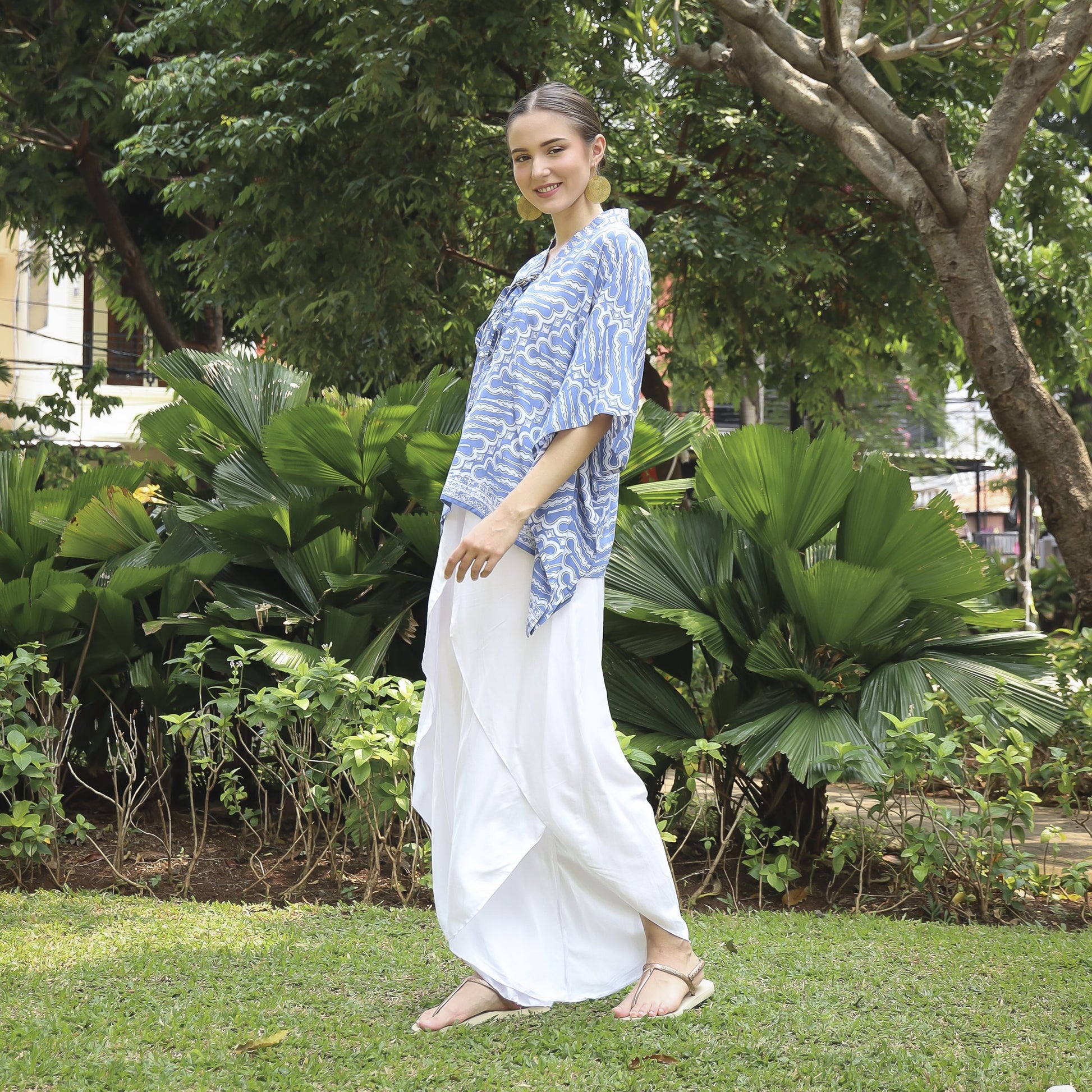 A person stands on grass, smiling and wearing a blue patterned top and white flowing pants, with lush greenery and trees in the background.
