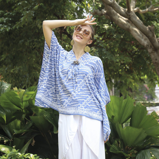 A person wearing a comfortable batik top for women and white pants smiles while shielding their eyes from the sun. They are standing outdoors in front of lush green foliage and trees.