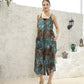 A woman in a patterned teal and brown dress, resembling the effortless style of a short jumpsuit, stands against a white textured wall. With her hands in her pockets and sandals on her feet, decorative pots add charm to the backdrop.