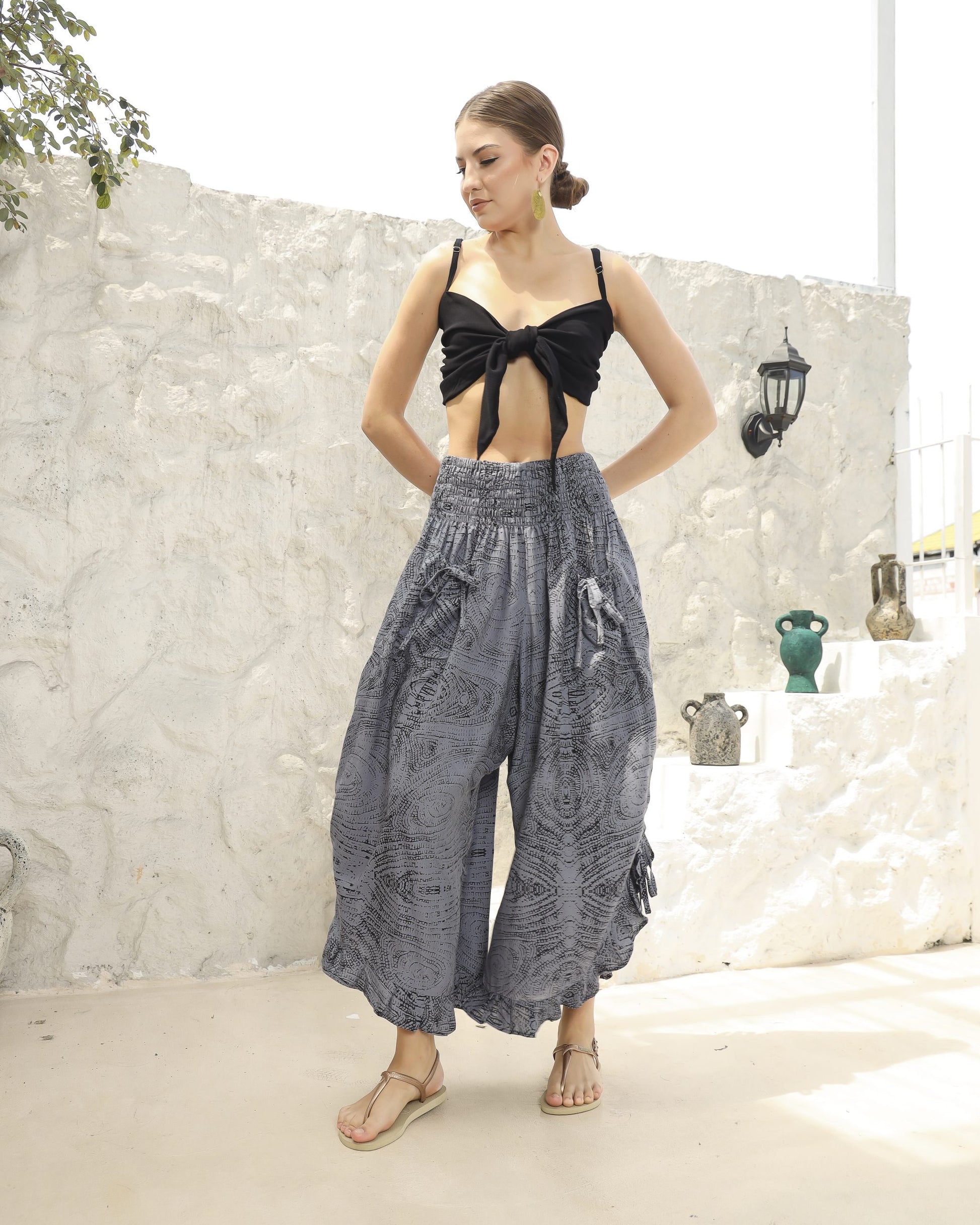 A woman stands outdoors against a textured white wall, wearing a black tie-front crop top and boho pants with a ruffle hem, paired with beige sandals. Her hair is pulled back as she gazes to the side. Decorative pots are arranged on a shelf nearby.