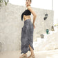 A woman stands confidently on a light-colored outdoor patio wearing a black bikini top with a front tie, gray patterned harem pants, and beige sandals. Her hair is styled in a ponytail, and she accessorizes with large hoop earrings. Decor is visible in the background.