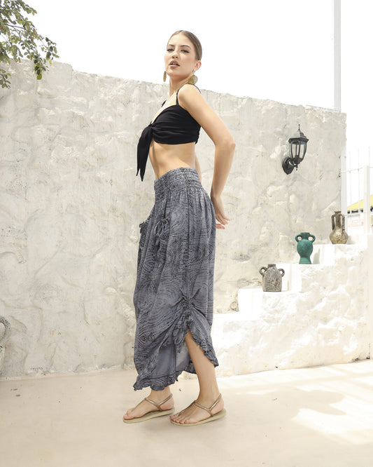 A woman stands confidently on a light-colored outdoor patio wearing a black bikini top with a front tie, gray patterned harem pants, and beige sandals. Her hair is styled in a ponytail, and she accessorizes with large hoop earrings. Decor is visible in the background.