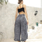 A person with their back to the camera stands outdoors against a textured white wall, wearing a black crop top that resembles a bikini top and patterned gray wide-leg pants with beige sandals. Decorative jugs are positioned on the stepped platform beside them.