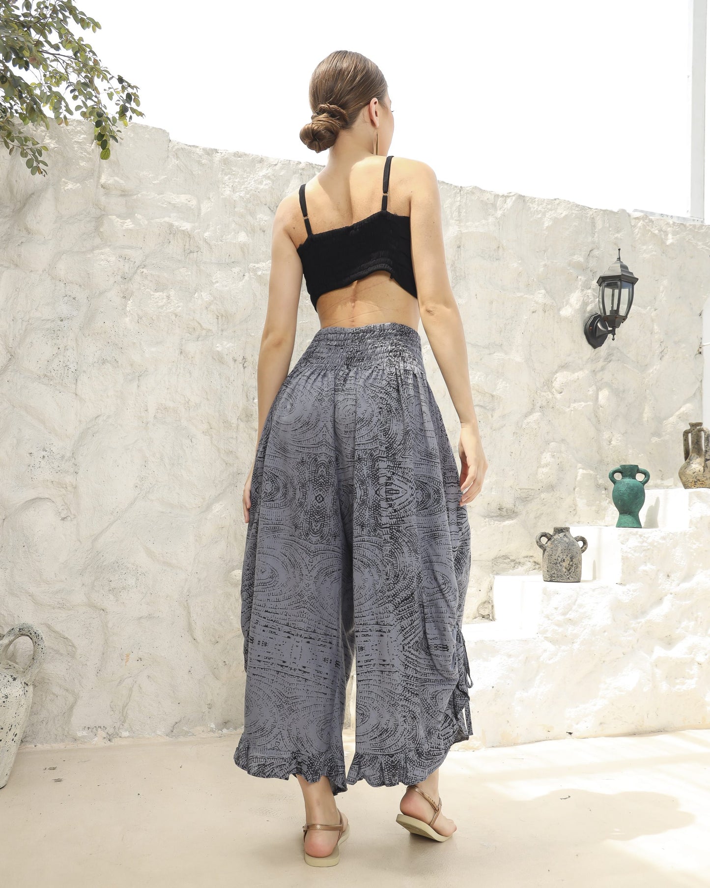 A person with their back to the camera stands outdoors against a textured white wall, wearing a black crop top that resembles a bikini top and patterned gray wide-leg pants with beige sandals. Decorative jugs are positioned on the stepped platform beside them.