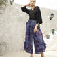 A woman stands confidently against a white stone wall, wearing a black top paired with boho pants featuring long, flowing blue-purple patterns. Her hands rest on her hips, and she completes the look with stylish sandals. Decorative pots adorn the steps beside her.