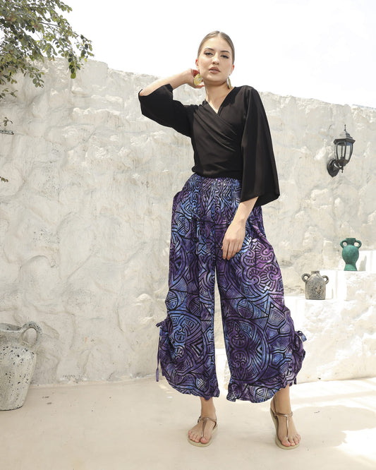 A woman stands confidently against a white stone wall, wearing a black top paired with boho pants featuring long, flowing blue-purple patterns. Her hands rest on her hips, and she completes the look with stylish sandals. Decorative pots adorn the steps beside her.