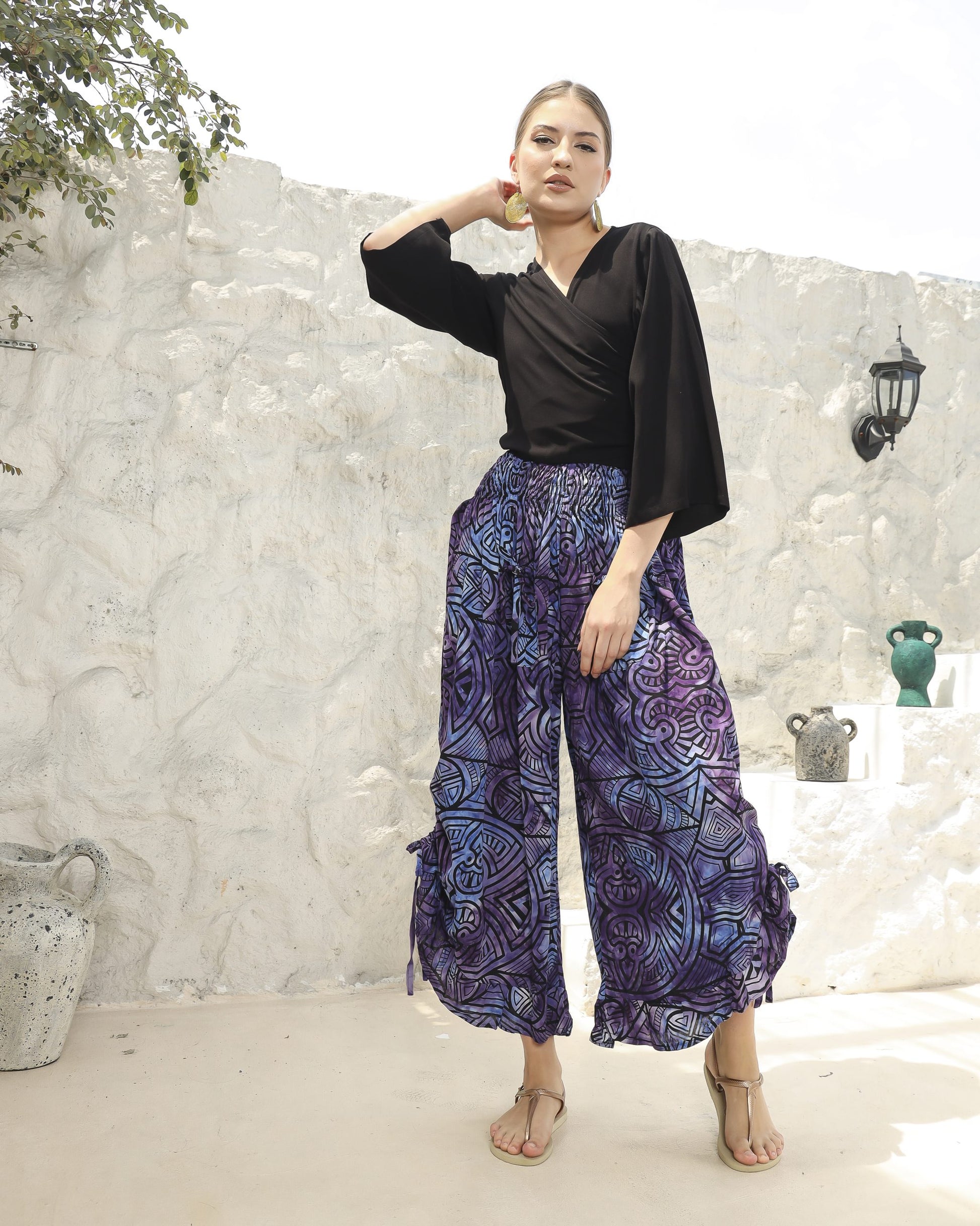 A woman stands confidently against a white stone wall, wearing a black top paired with boho pants featuring long, flowing blue-purple patterns. Her hands rest on her hips, and she completes the look with stylish sandals. Decorative pots adorn the steps beside her.
