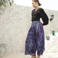A woman stands confidently in front of a textured white wall, wearing a chic black boho top with 3/4 sleeves and purple-patterned wide-leg pants. She completes her look with sandals and a sleek hairdo, one hand poised on her hip.