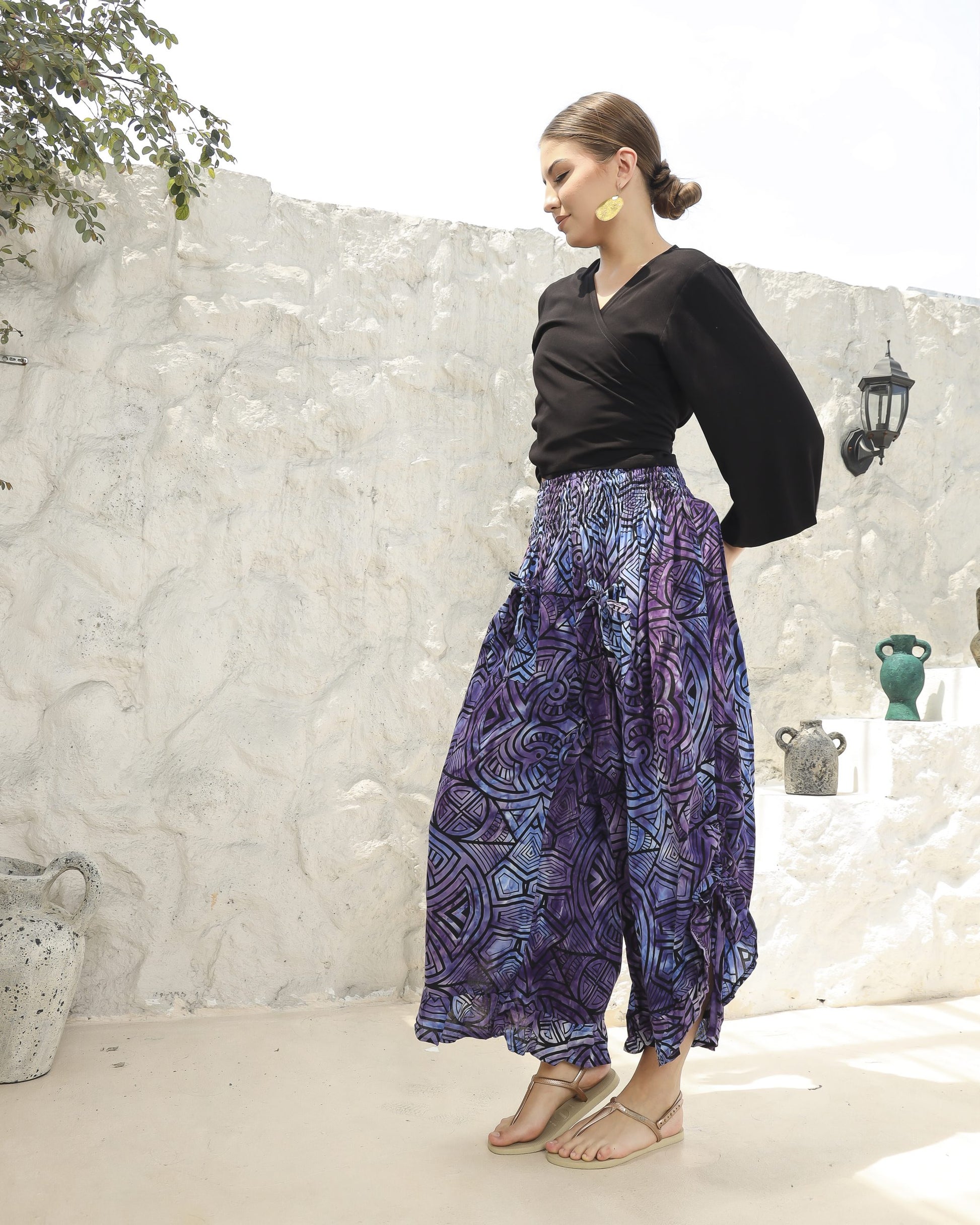 A woman stands confidently in front of a textured white wall, wearing a chic black boho top with 3/4 sleeves and purple-patterned wide-leg pants. She completes her look with sandals and a sleek hairdo, one hand poised on her hip.