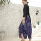 A woman stands outdoors against a textured white wall, wearing a black boho top with 3/4 sleeves and flowing purple patterned pants, paired with sandals. Decorative pots, a wall lamp, and a leafy branch enhance the serene backdrop.