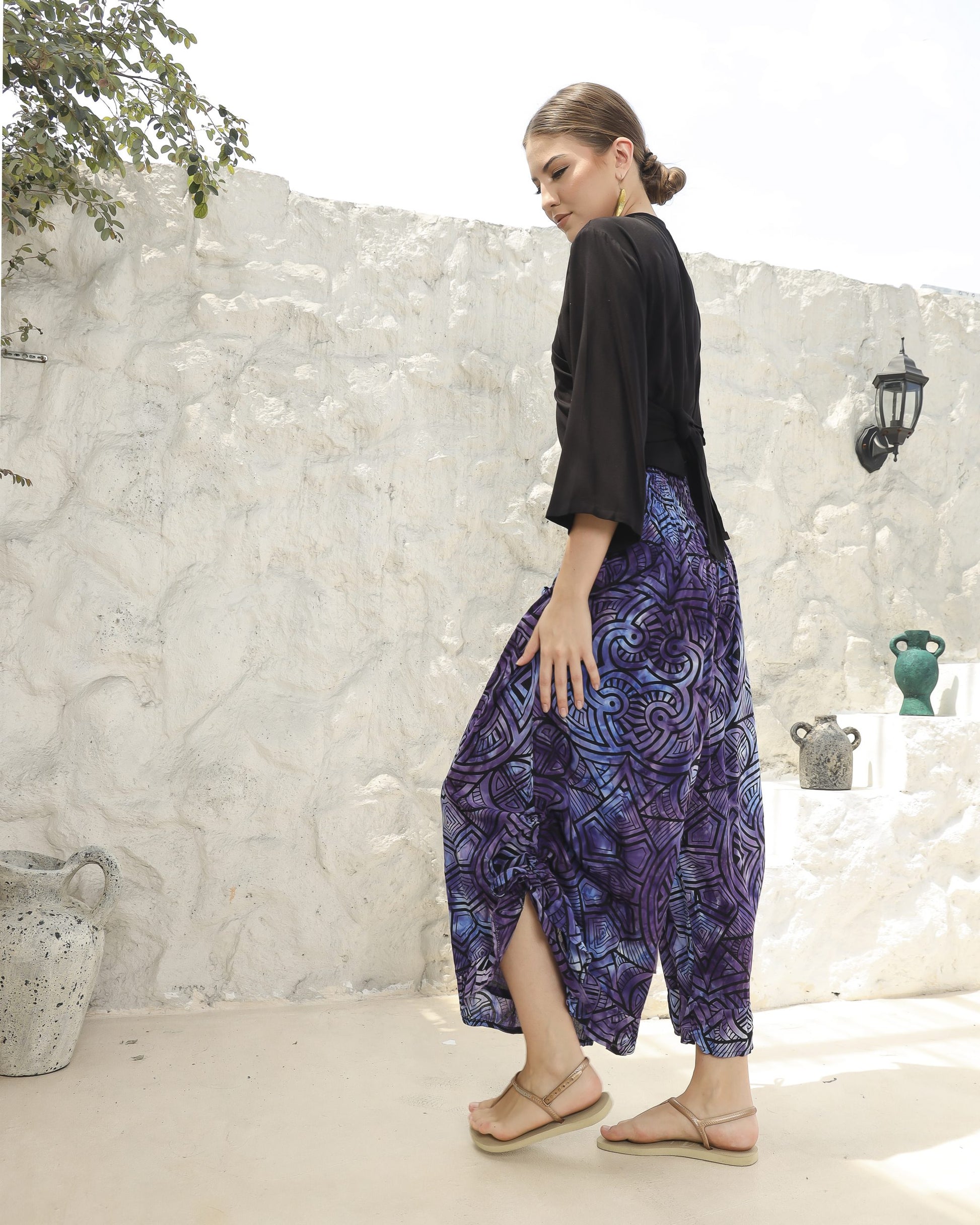 A woman stands outdoors against a textured white wall, wearing a black boho top with 3/4 sleeves and flowing purple patterned pants, paired with sandals. Decorative pots, a wall lamp, and a leafy branch enhance the serene backdrop.