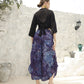 A woman stands outdoors with her back to the camera, wearing a boho top with 3/4 sleeves and blue, patterned pants. Her hair is tied back, and she is in sandals. The background features a white stone wall and a few potted plants.