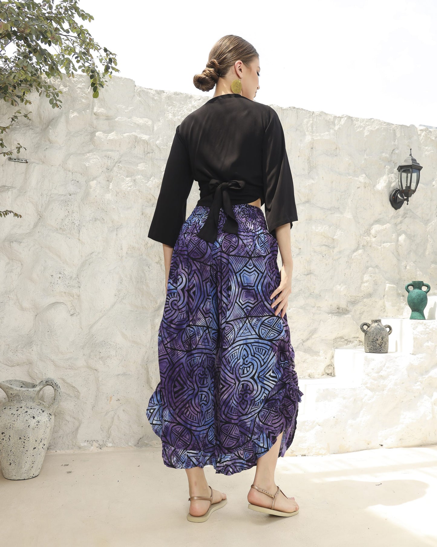 A woman stands outdoors with her back to the camera, wearing a boho top with 3/4 sleeves and blue, patterned pants. Her hair is tied back, and she is in sandals. The background features a white stone wall and a few potted plants.