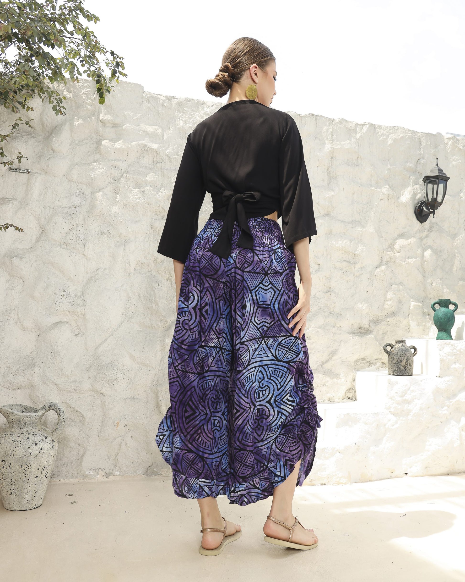 A woman stands outdoors with her back to the camera, wearing a boho top with 3/4 sleeves and blue, patterned pants. Her hair is tied back, and she is in sandals. The background features a white stone wall and a few potted plants.