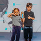 A young girl with a Barbie doll poses next to a boy giving a thumbs-up. They stand in front of a backdrop with Rinjanie text. The girl sports yoga pants for kids in purple, paired with a grey shirt, while the boy wears a black shirt and dark pants.