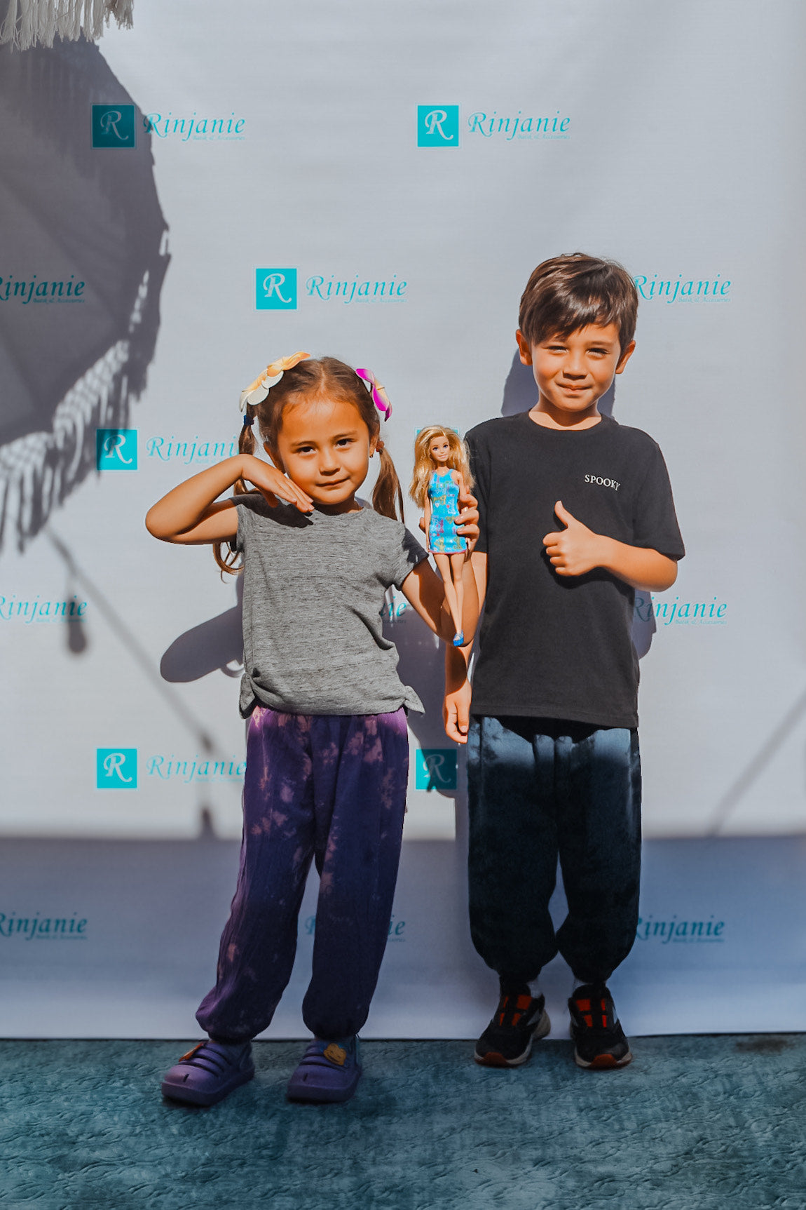 A young girl with a Barbie doll poses next to a boy giving a thumbs-up. They stand in front of a backdrop with Rinjanie text. The girl sports yoga pants for kids in purple, paired with a grey shirt, while the boy wears a black shirt and dark pants.