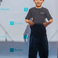 A young child stands on a turquoise carpet, smiling and giving two thumbs up. Sporting a black t-shirt with SPOOKY, dark yoga pants for kids, and sneakers, the child exudes joy. The background features a pattern with the word Rinjanie and a decorative R logo.