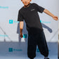 A young boy with dark hair is posing in front of a backdrop with the word Rinjanie repeated on it. He is wearing a black T-shirt with SPOOKY on it, dark pants, and sneakers. His right arm is bent, and he smiles slightly.