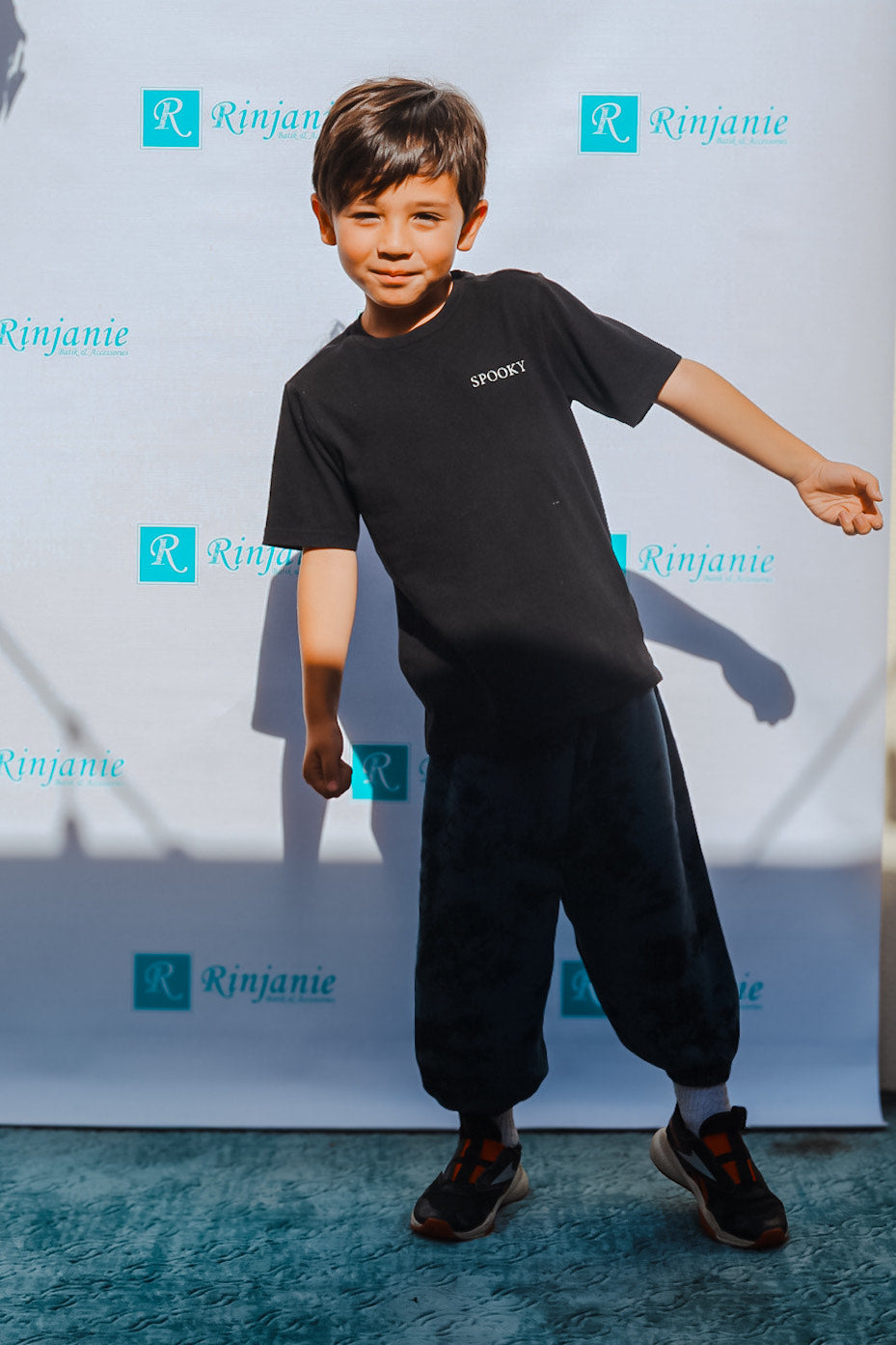 A young boy with dark hair is posing in front of a backdrop with the word Rinjanie repeated on it. He is wearing a black T-shirt with SPOOKY on it, dark pants, and sneakers. His right arm is bent, and he smiles slightly.