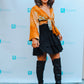 A person wearing a large brown hat, orange patterned top, black tiered skirt, and knee-high black boots stands in front of a backdrop with blue Rinjanie logos. They are smiling and posing for the camera.