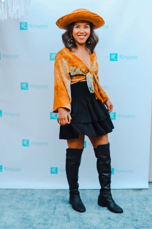 A person wearing a large brown hat, orange patterned top, black tiered skirt, and knee-high black boots stands in front of a backdrop with blue Rinjanie logos. They are smiling and posing for the camera.