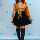 A woman in an orange tie-dye top, a mini skirt with layers, and black knee-high boots poses in front of a backdrop. She wears a wide-brimmed hat and smiles at the camera, while the backdrop proudly displays a repeated logo.