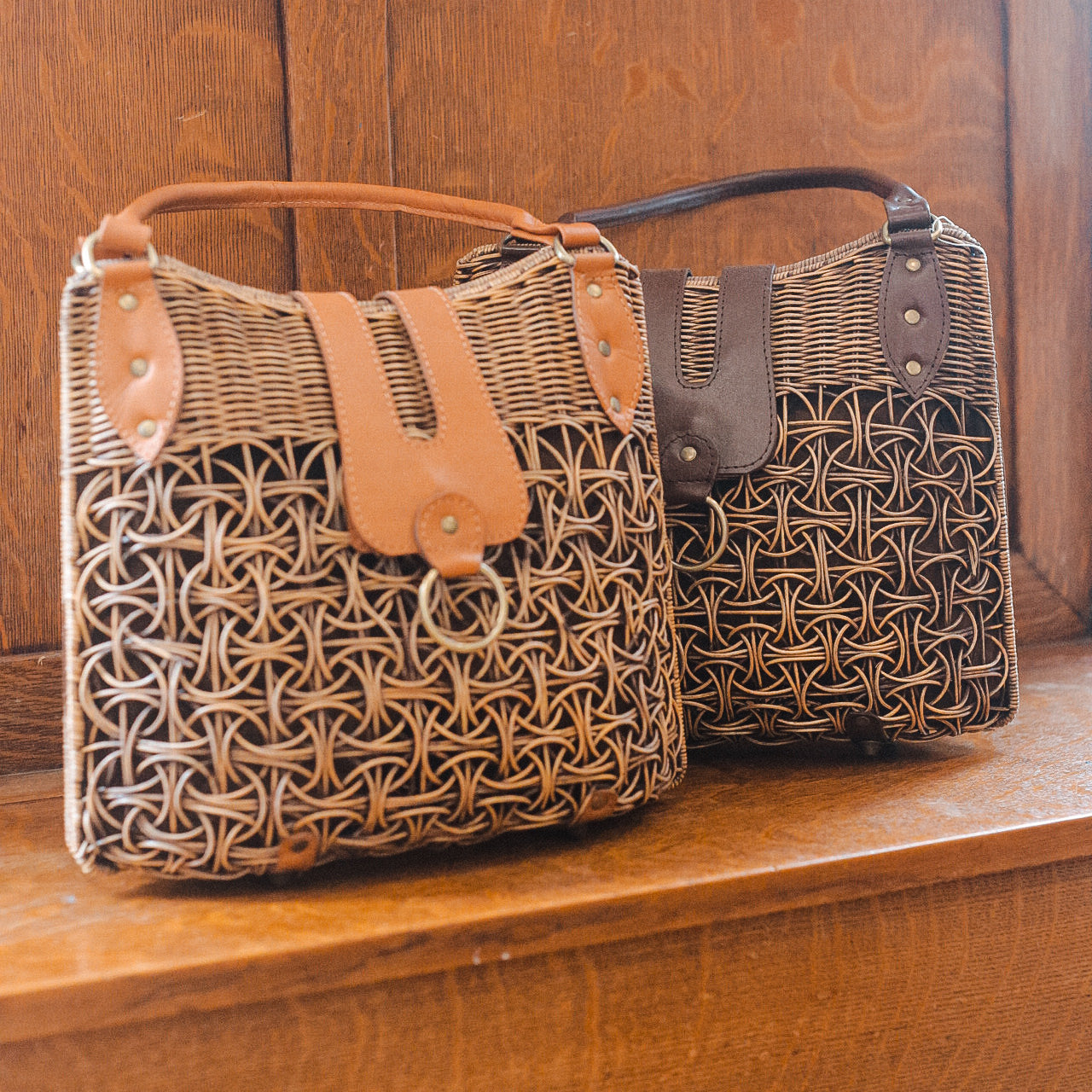 Two woven handbags, reminiscent of a large rattan purse, rest on a wooden surface with intricate patterns. Each bag features a leather flap closure, one in tan and the other in dark brown. The bags flaunt matching leather handles against a backdrop of dark wood paneling.