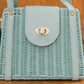 One of our light blue small rattan purses with a rectangular shape, featuring a flap closure secured by a gold twist lock. The handbag has a matching light blue strap and brass-colored accents, set against a wooden background.