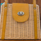 A wicker handbag with tan leather accents, featuring a front flap secured by a metal twist-lock clasp. The design is structured and has two leather handles.