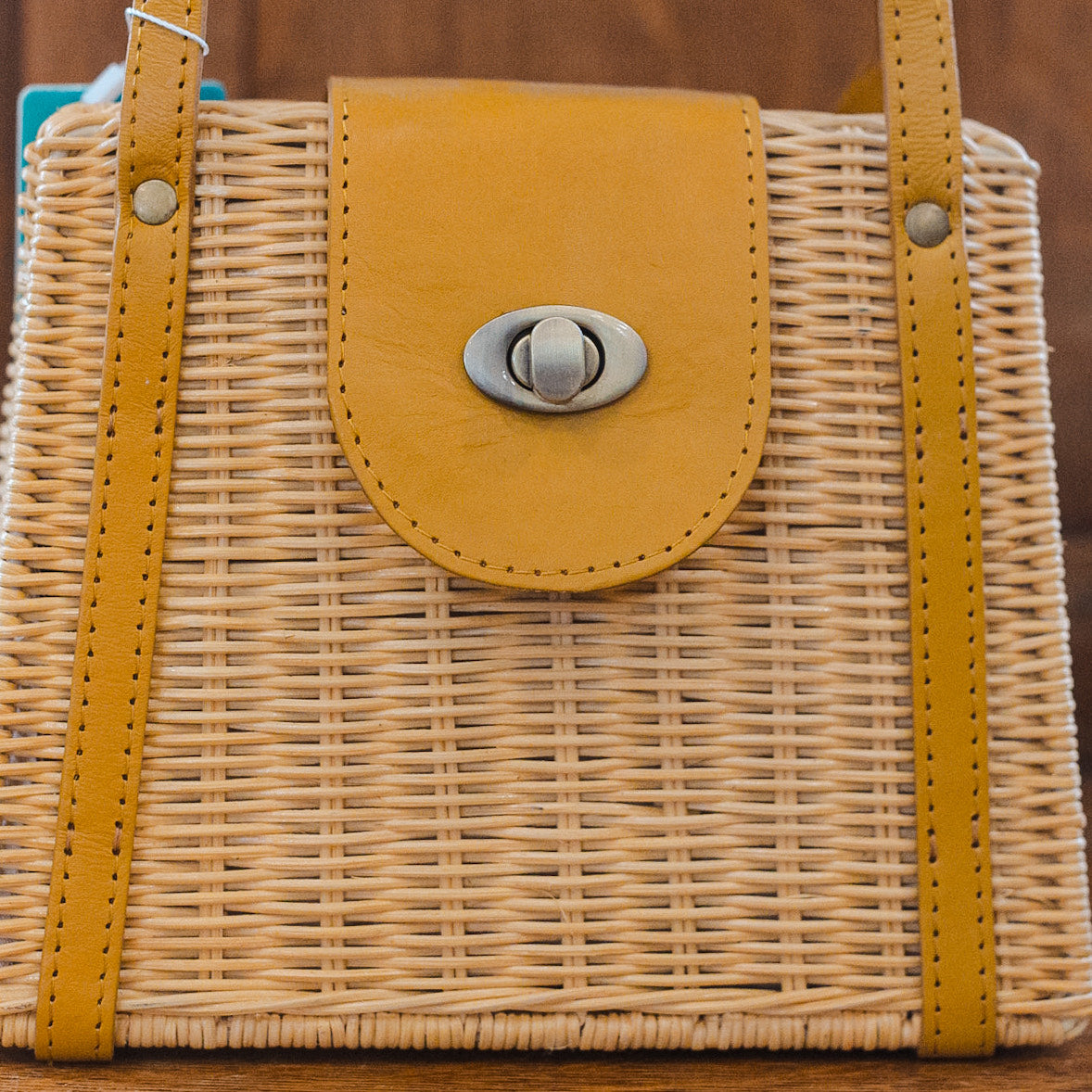 A wicker handbag with tan leather accents, featuring a front flap secured by a metal twist-lock clasp. The design is structured and has two leather handles.