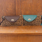 Two woven straw clutch purses for women, each with a leather flap, are displayed on a wooden surface. One bag features a brown flap, the other teal. Both boast circular metal clasps and intricate wicker patterns.