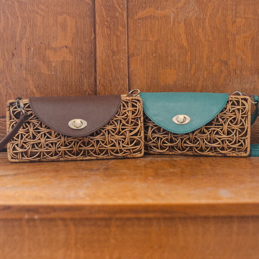 Two woven straw clutch purses for women, each with a leather flap, are displayed on a wooden surface. One bag features a brown flap, the other teal. Both boast circular metal clasps and intricate wicker patterns.