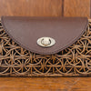 A woven clutch purse for women showcases a circular rattan pattern and a dark brown leather flap, complemented by a metallic clasp at the front. The bag rests elegantly on a wooden surface background.