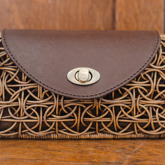 A woven clutch purse for women showcases a circular rattan pattern and a dark brown leather flap, complemented by a metallic clasp at the front. The bag rests elegantly on a wooden surface background.