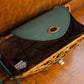 A small woven rattan purse with a green flap is open, revealing a dark brown interior. It rests on a wooden surface, with visible metal fittings and a tag inside.