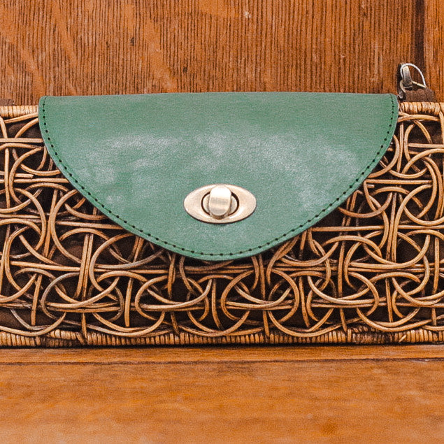 A clutch purse made of woven rattan with a green leather flap and silver clasp, placed against a wood background. A clutch purse made of woven rattan with a green leather flap and silver clasp, placed against a wood background.