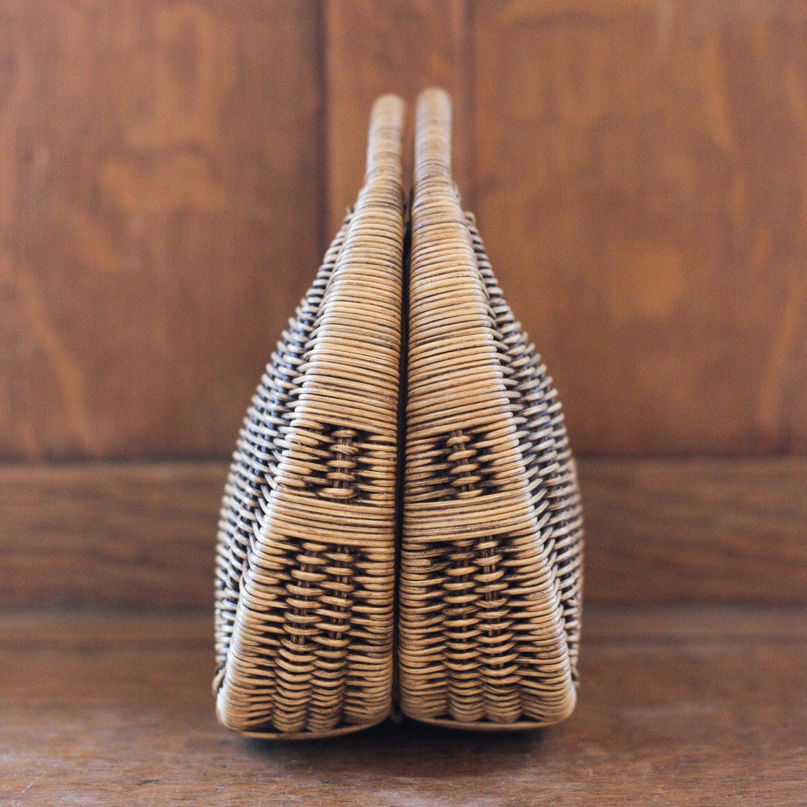 A woven wicker handbag standing upright on a wooden surface against a wooden background. The bag has a symmetrical design with a tightly knit pattern, featuring natural brown tones.