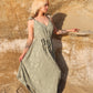 A woman with blonde hair stands against a sandy rock wall, wearing a green patterned long jumpsuit. She has tattoos on her arms and holds the edge of her outfit while looking to the side. Sunlight casts a warm glow on the scene. A woman with blonde hair stands against a sandy rock wall, wearing a green patterned long jumpsuit. She has tattoos on her arms and holds the edge of her outfit while looking to the side. Sunlight casts a warm glow on the scene.