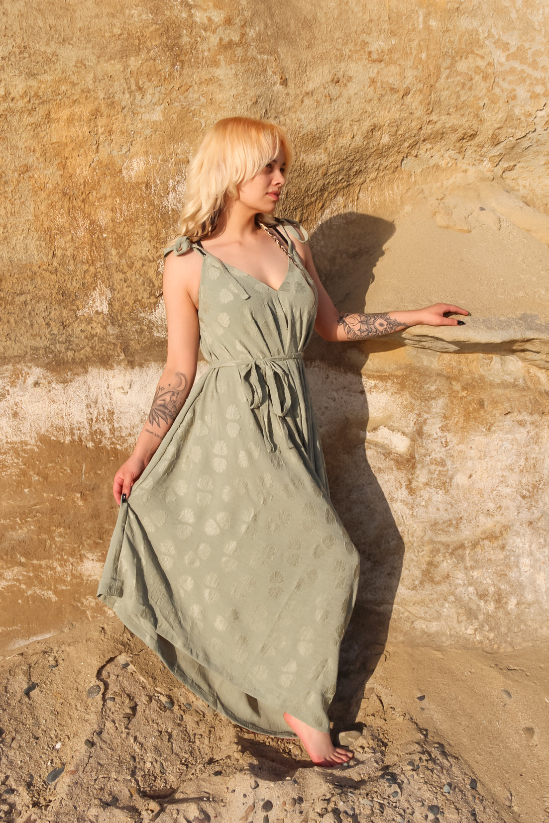 A woman with blonde hair stands against a sandy rock wall, wearing a green patterned long jumpsuit. She has tattoos on her arms and holds the edge of her outfit while looking to the side. Sunlight casts a warm glow on the scene. A woman with blonde hair stands against a sandy rock wall, wearing a green patterned long jumpsuit. She has tattoos on her arms and holds the edge of her outfit while looking to the side. Sunlight casts a warm glow on the scene.