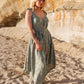 A woman with blonde hair stands barefoot on a sandy beach, wearing a green patterned long jumpsuit. She poses confidently against the backdrop of a sunlit, rocky cliff, creating a natural and serene atmosphere.
