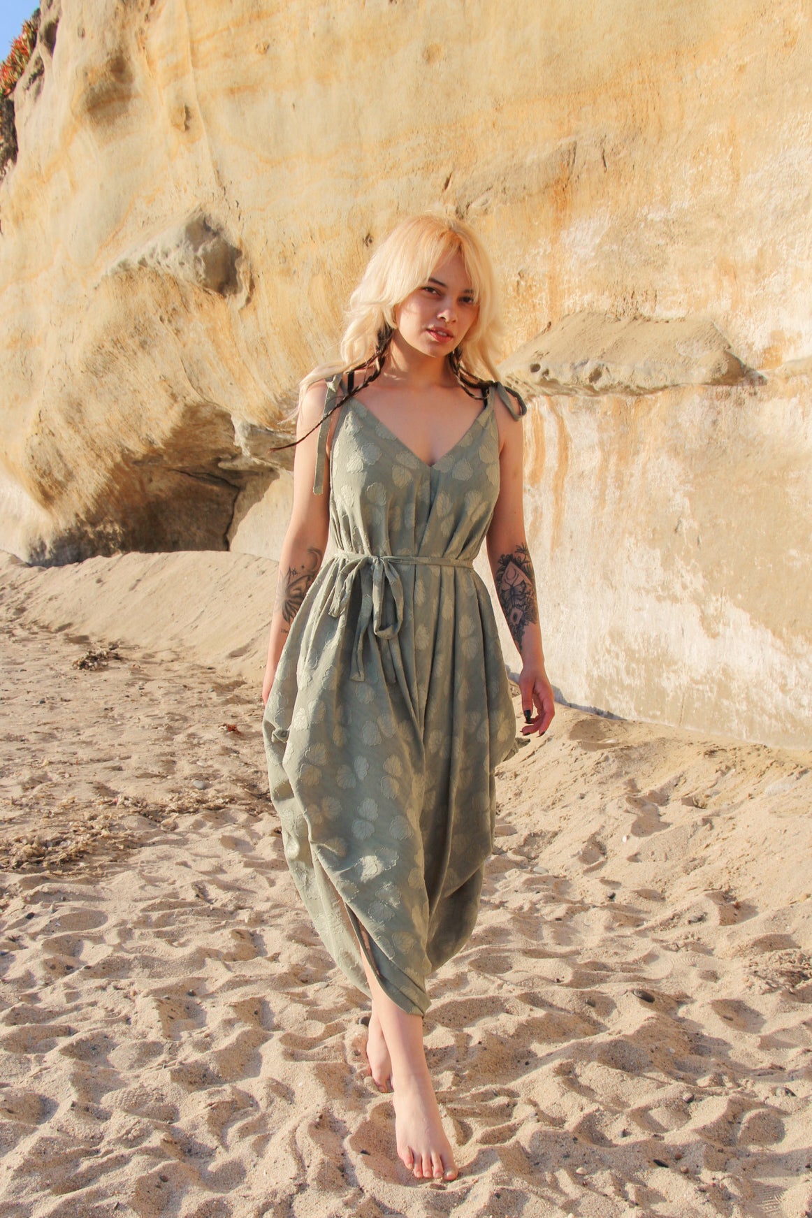 A person with blonde hair walks barefoot on a sandy beach, wearing a flowing green dress that resembles the elegance of a long jumpsuit. The backdrop features a large rock formation with a light brown and beige texture.